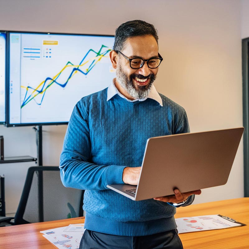 Happy-middle-aged-businessman-using-a-laptop-with-a-digital-growth-chart-displayed-on-a-screen-in-a-modern-office-setting.