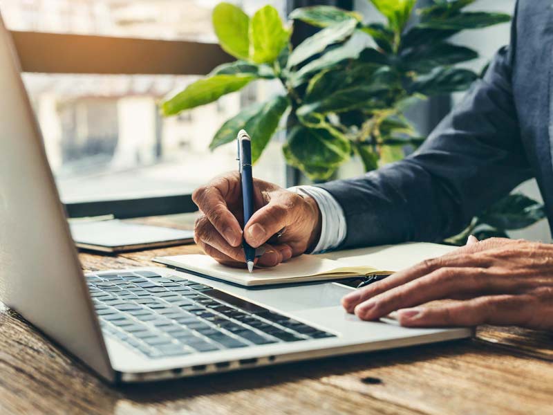 Close-up-of-a-man-writing-notes-in-a-notebook-next-to-a-laptop-on-a-wooden-desk,-with-green-plants-in-the-background.