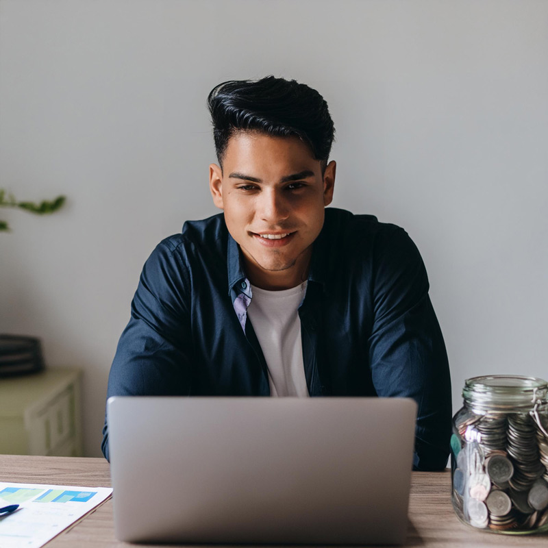 Young-smiling-man-sitting-at-a-desk-using-a-laptop-with-a-jar-full-of-coins,-illustrating-personal-finance-management.